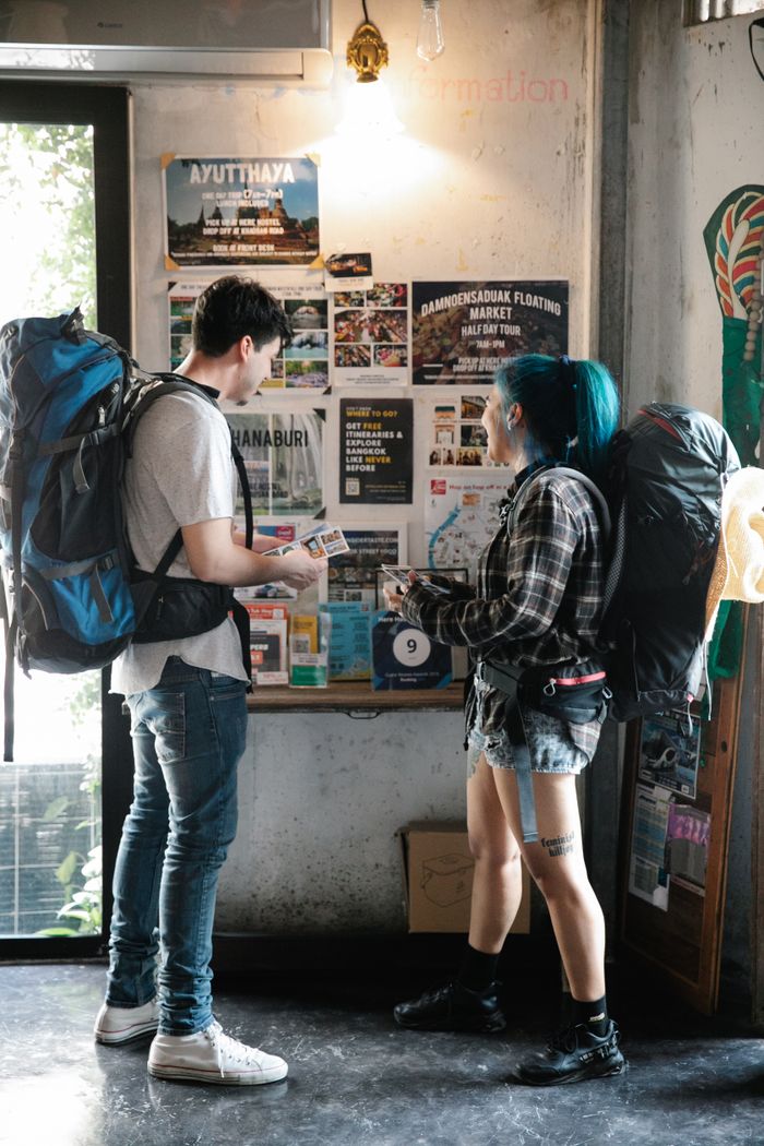 Side view of young anonymous couple of tourists in casual clothes and with backpacks looking at information board in hotel